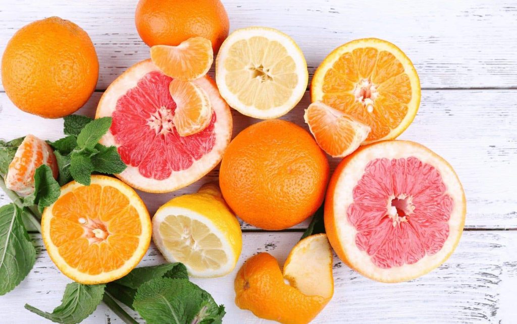 A variety of citrus fruits are displayed on a white wooden surface. Whole and halved oranges, lemons, and grapefruits are mixed with peeled segments. There are also green leaves scattered among the fruits, showcasing the health benefits of citrus in a vibrant array. MyFitnessPal Blog