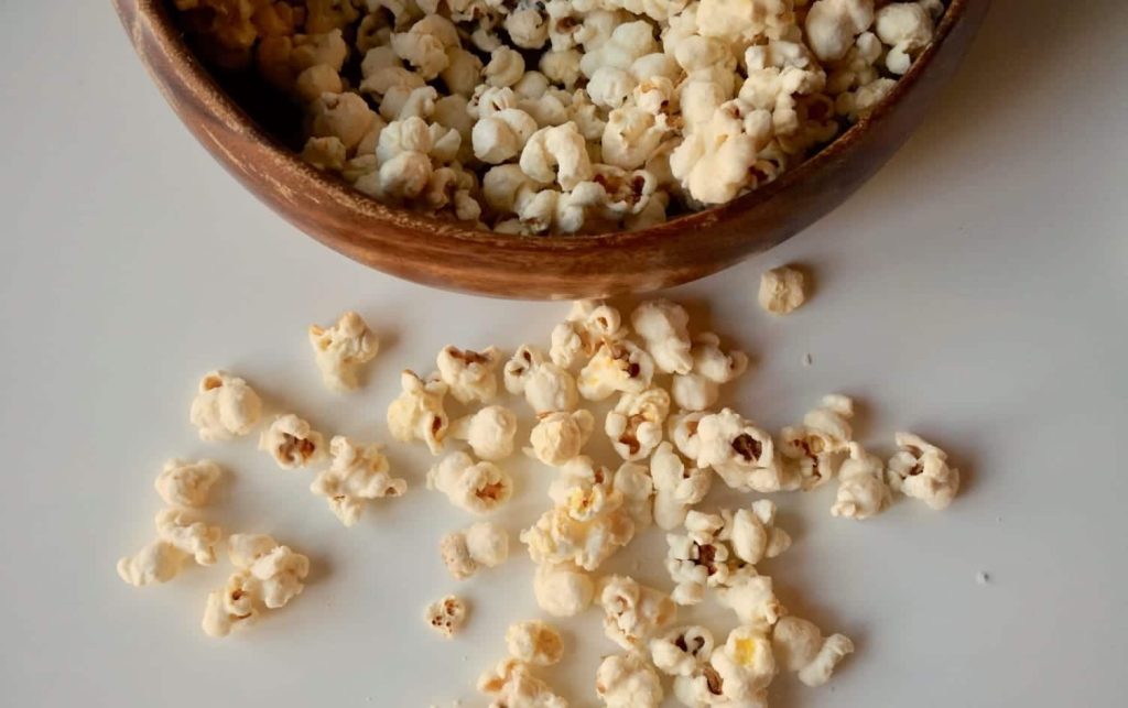 A wooden bowl filled with popcorn sits on a white surface. Some of the fluffy popcorn has spilled out, scattering around it. The popcorn appears to be plain, without any visible toppings or seasoning. MyFitnessPal Blog