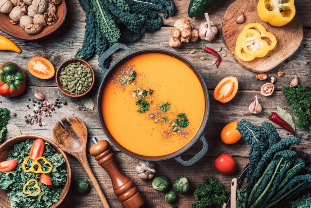 Pumpkin soup surrounded by fresh vegetables, kale, peppers, and cooking ingredients on wooden table