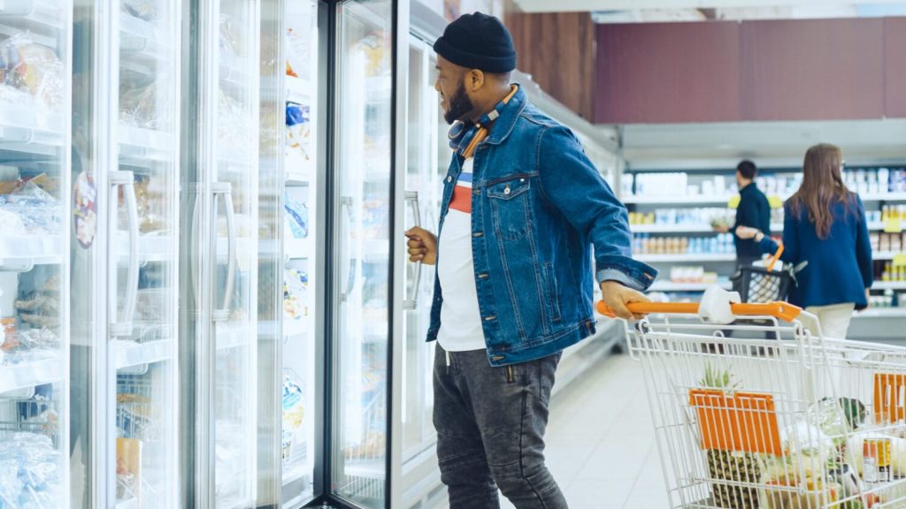 Man in denim jacket shopping for frozen vegetables in supermarket freezer aisle
