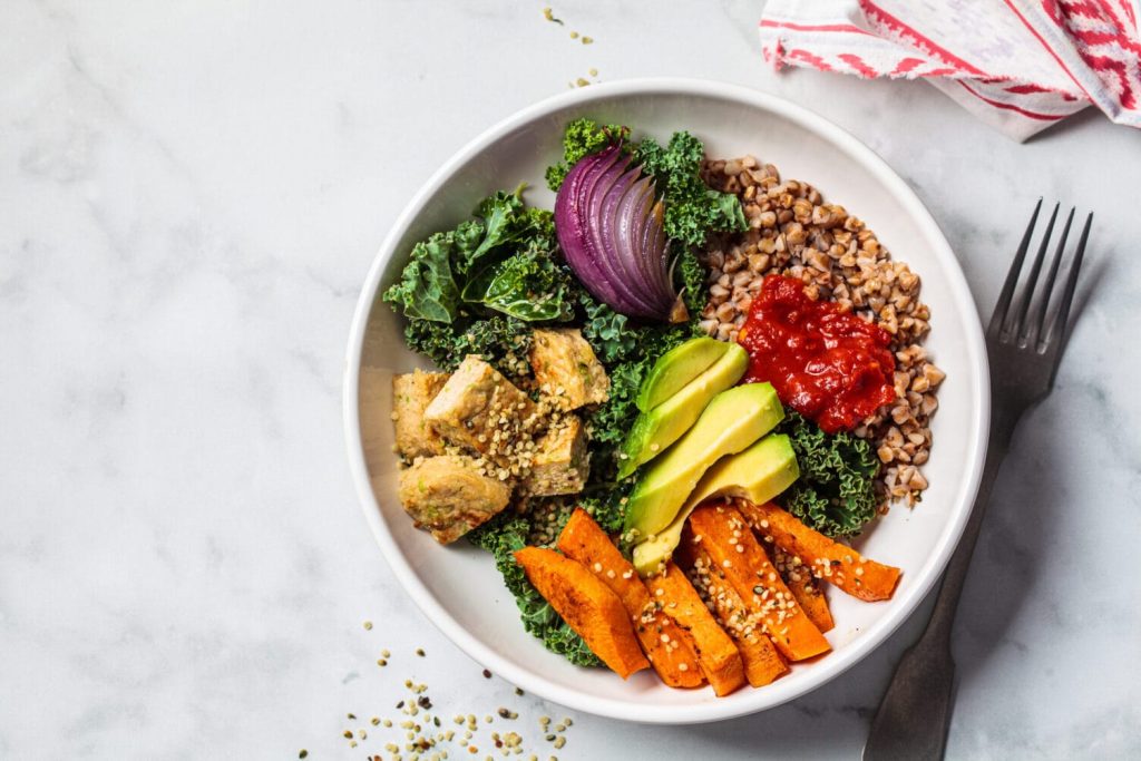 Colorful vegan buddha bowl with kale, tofu, avocado, sweet potato, buckwheat and red onion on white plate