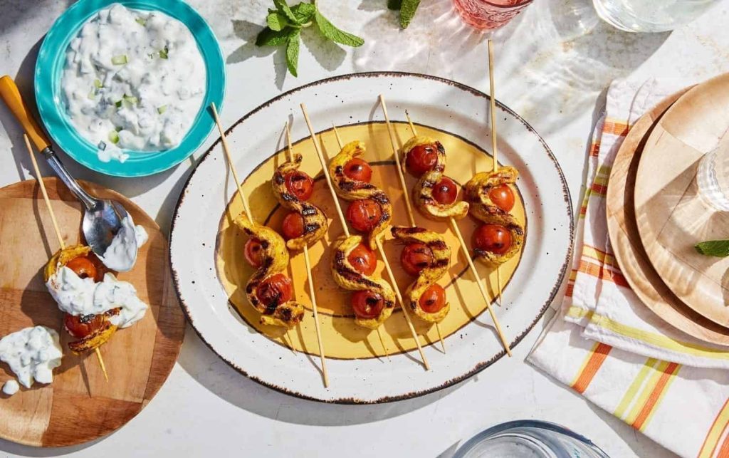 Curried Chicken Skewers With Raita Grilled shrimp and cherry tomato skewers, alongside chicken skewers, arranged on a yellow oval dish, accompanied by a bowl of tzatziki sauce. A wooden plate with a skewer and sauce sits nearby, along with a napkin holder featuring plaid napkins and a partly visible glass of water, all set on a white surface. MyFitnessPal Blog