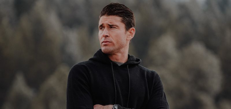 Ben Greenfield’s Weekly Roundup: September 11 Portrait of a Ben Greenfield with short dark hair wearing a black hoodie, arms crossed, standing outdoors with a blurred backdrop of evergreen trees. He looks off to the side with a serious expression.